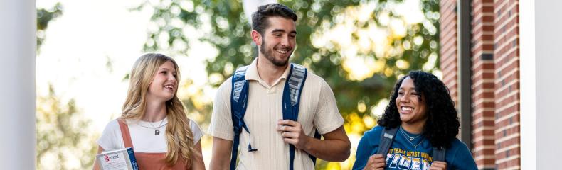 Three college students walk and talk together on campus, smiling and carrying backpacks and books.