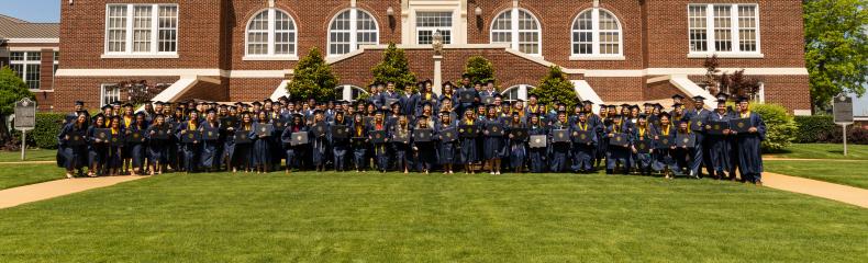 A large group of graduates in graduation regalia stand in front of ETBU's Historic Marshall Hall