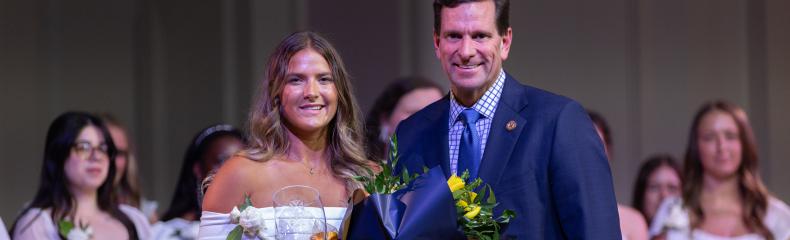 2 people in formal clothing smiling in front of a large group of people in white clothing behind them