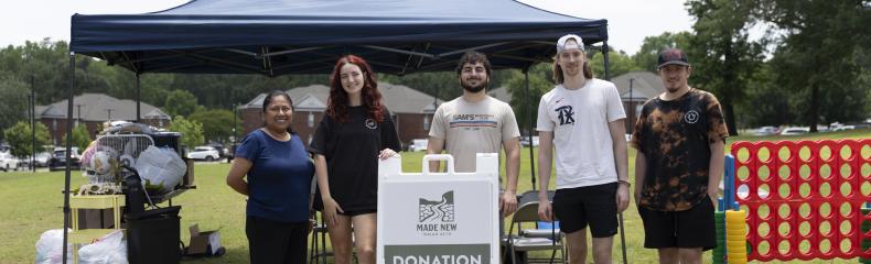 Five people stand in front of a blue canopy tent at a donation drop-off event, with a sign reading "Donation Drop-Off" placed in the center.