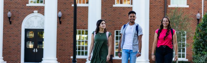 Two females and one male ETBU student walking down a side walk in front of a red brick building with white columns in the background.