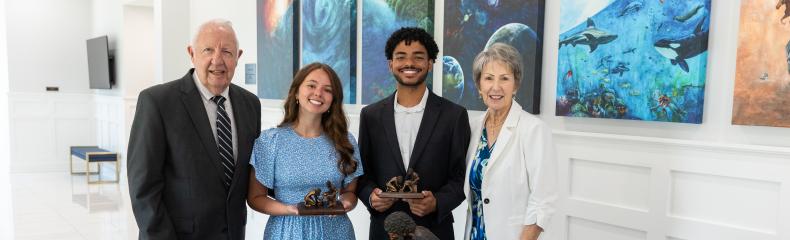 Bob and Gayle Riley stand on either side of a male and female student holding awards behind a table inside a building with white walls.