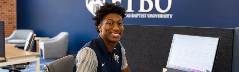 A man sitting at a table with a computer on it and the ETBU logo in the background