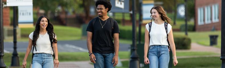 Three students walk down a campus sidewalk, smiling at each other.