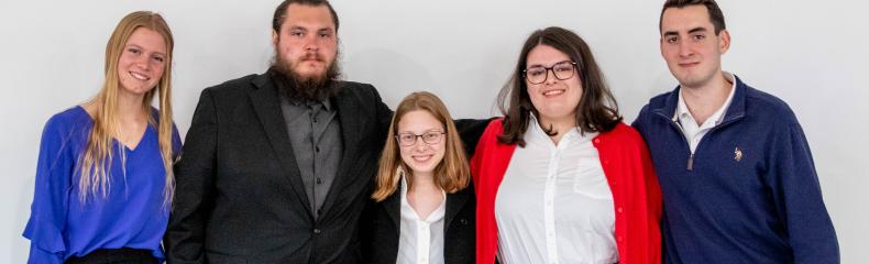 Five students stand side by side in professional attire and pose for a photo in front of a white wall.