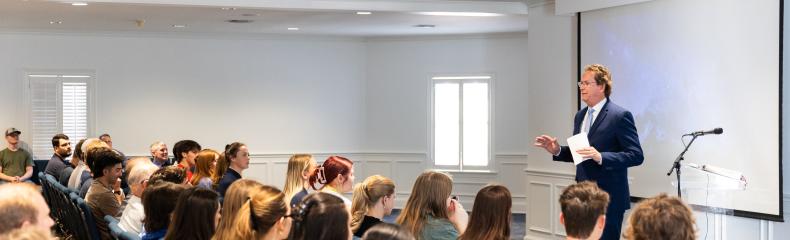 Rows of students are seated in blue chairs listening to a speaker who is standing in front of a projector screen.
