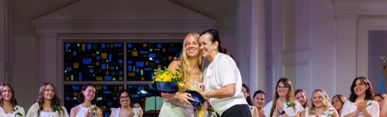 A smiling woman hands another smiling woman a bouquet of flowers as she accepts an award on stage. Several women who are wearing white dresses and applauding are shown behind the pair in the center of the stage.