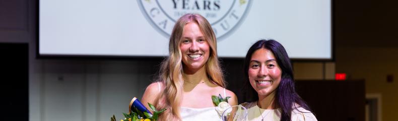 Two girls stand in white dresses. One is holding a bouquet of flowers and there is an event slide shown on a projector screen behind them.