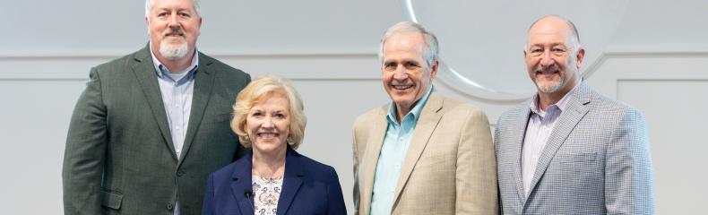 Four people stand together for a photo, each wearing a blazer and smiling