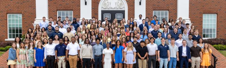 A large group of more than 100 students stands on an exterior staircase showing their class rings