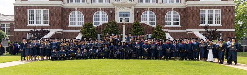 A group of graduates in navy regalia standing in front of a red brick building smiling.