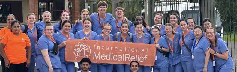 A group of students in blue scrubs stand with a International Medical Relief orange sign in front of a scenic backdrop.