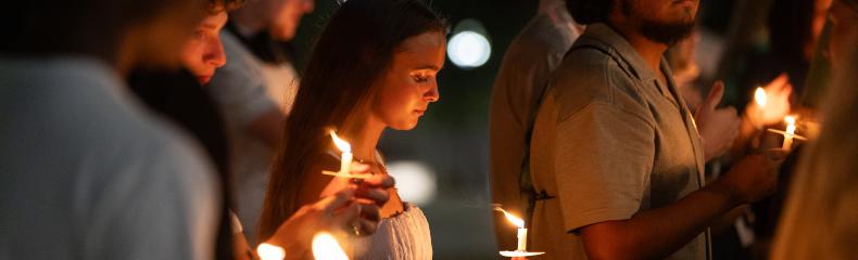Photograph of a candlelight vigil held outdoors at night, showing a group of people holding lit candles with solemn expressions. Warm candlelight illuminates faces against a dark background, highlighting a sense of unity and remembrance.